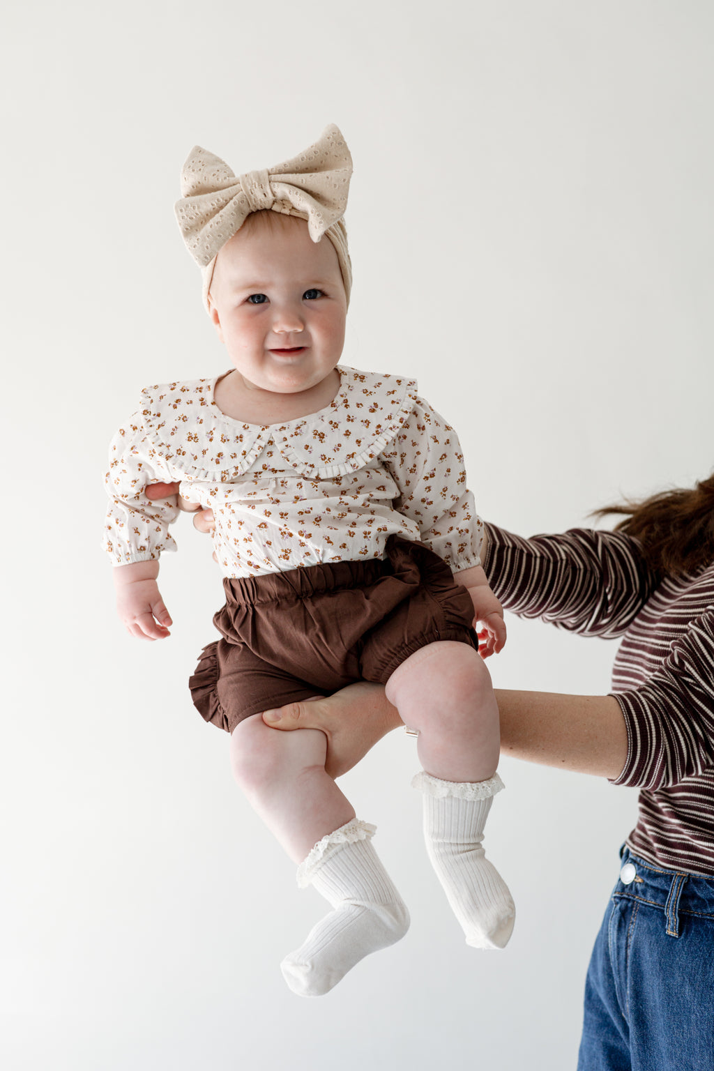Baby being held up by her mom. She is wearing a bow, a romper and brown short pants.