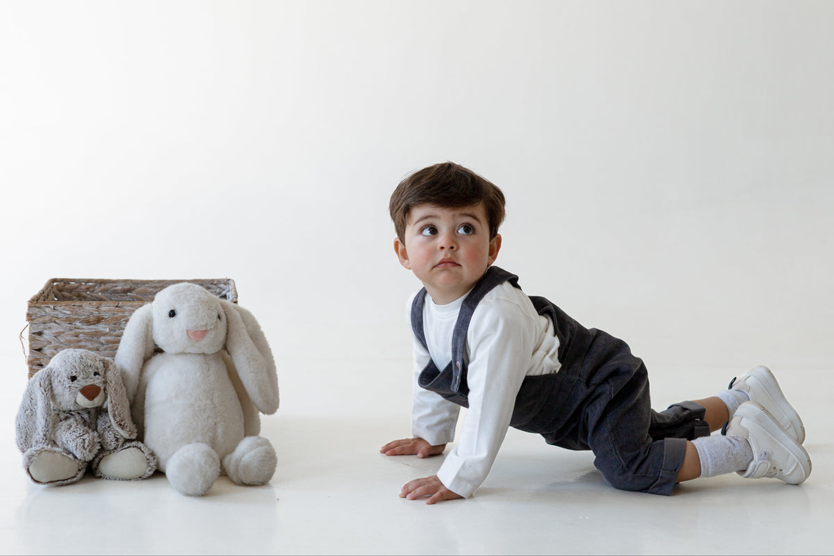 A toddler boy in a white long-sleeve shirt and dark navy overalls crawls on a white studio floor next to a brown basket and two plush bunny toys.