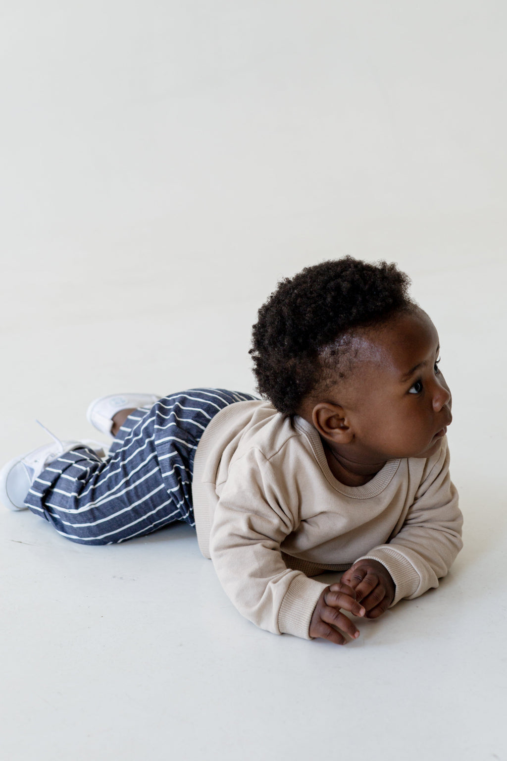 A baby lying on their stomach, wearing a beige long-sleeve top and navy blue and white striped pants, against a plain off-white background.
