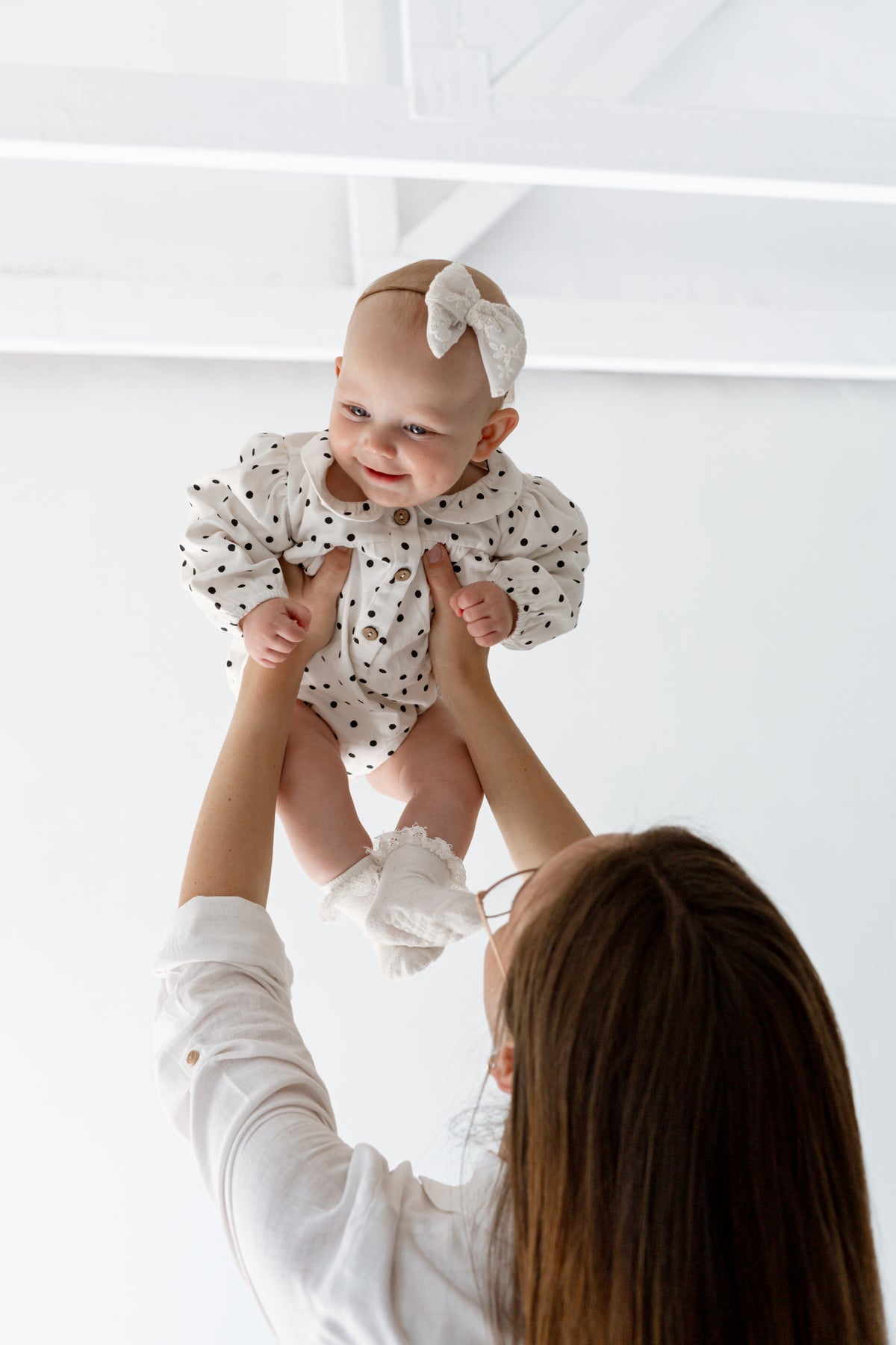 A baby wearing a white long-sleeve romper with black polka dots and a matching bow headband is being held up by an adult against a bright white background.