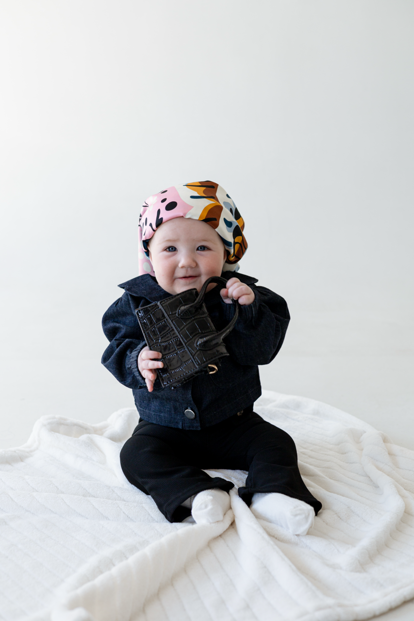 Baby wearing a denim shacket and holding a black purse with a bandana on her head.