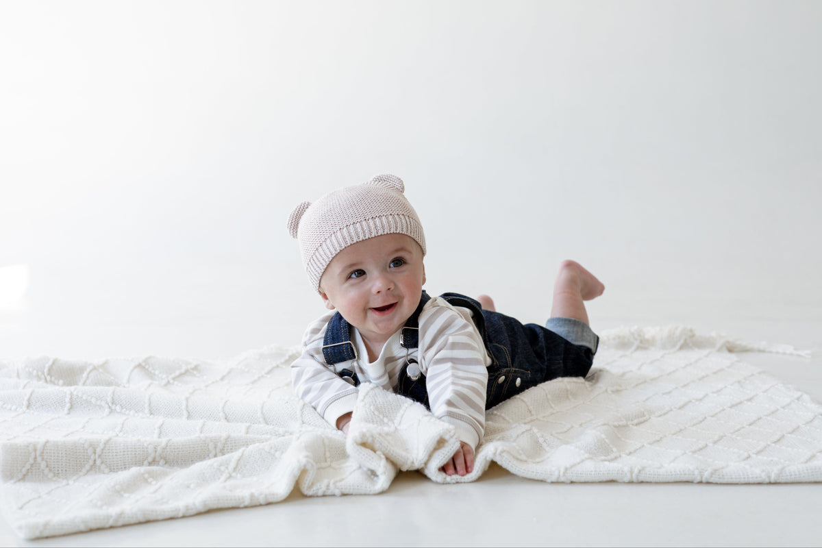 A baby lying on a white textured blanket, wearing a light pink knit beanie with ear details, a white and gray striped long-sleeve shirt, and dark blue denim overalls.