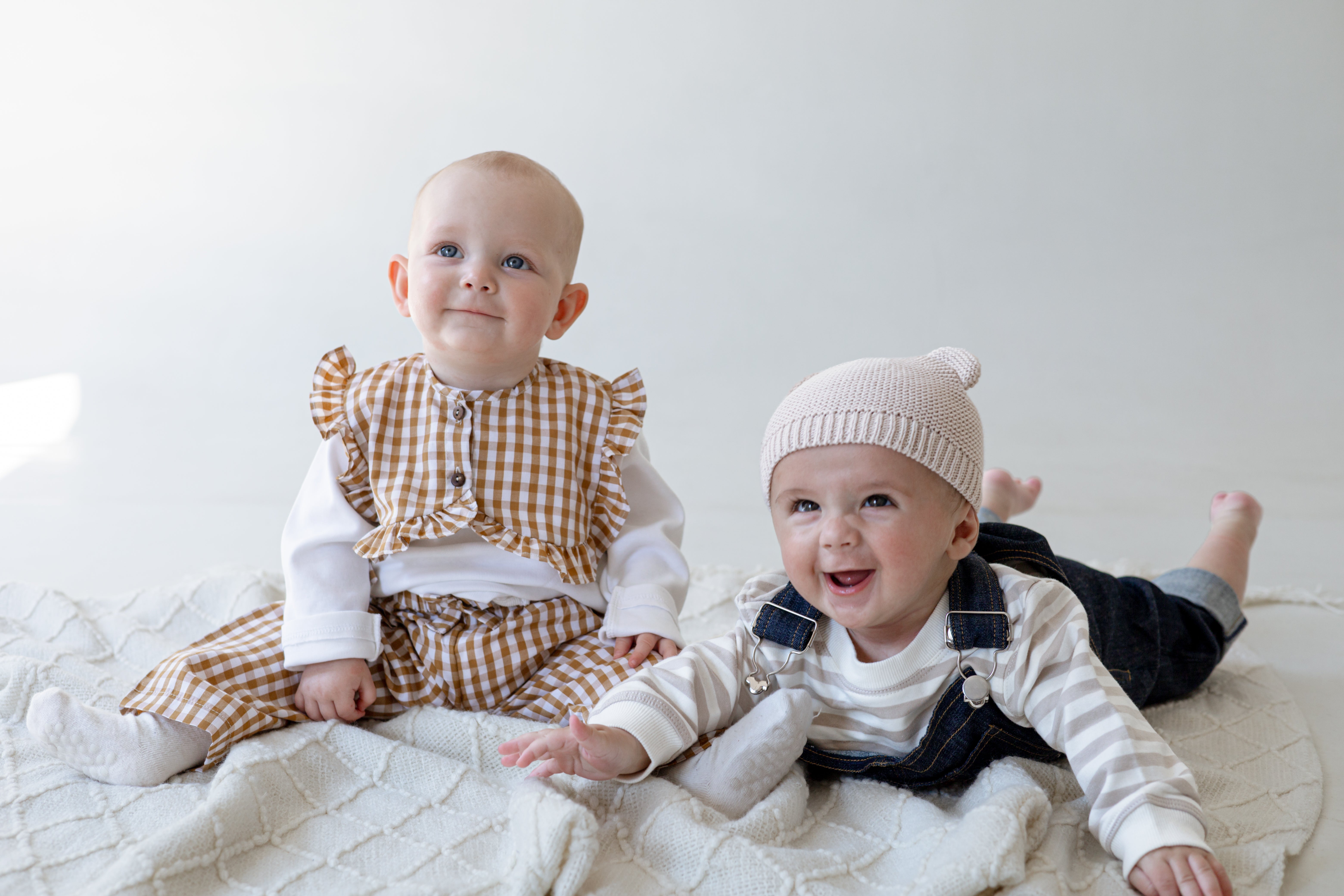 Two babies on the floor. One sitting and one laying on his stomach on the white blanket.